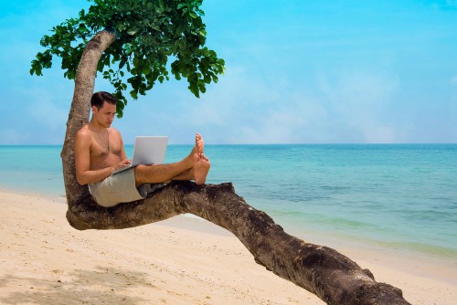 Красивый парень с ноутбуком в руках сидит на дереве на тропическом острове